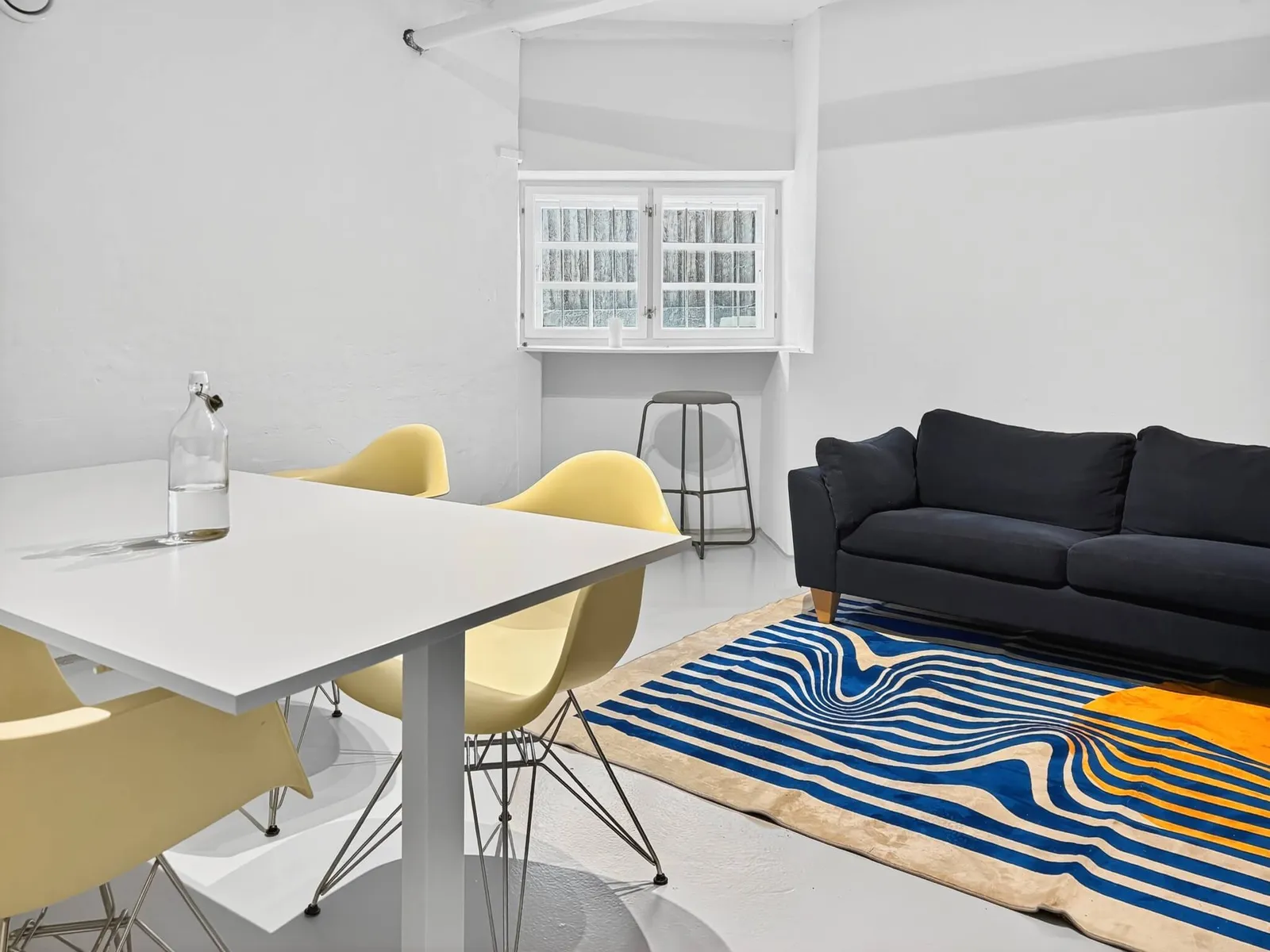 Hjørne med mørkeblå sofa, hvidt bord, gule designerstole og blåt bølgemønstret tæppe